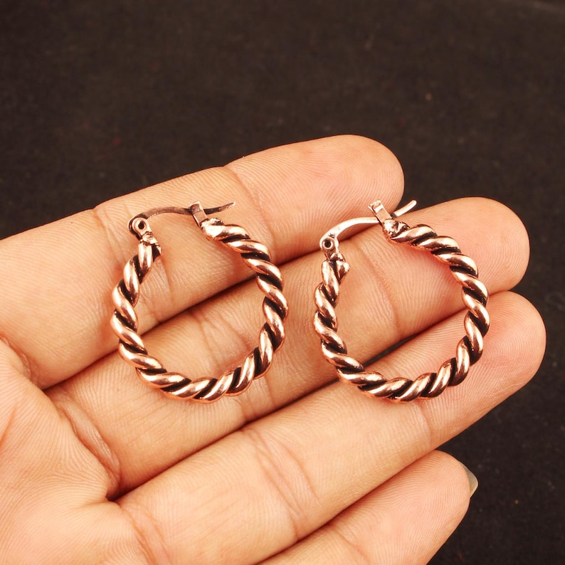 May include: A pair of copper-toned hoop earrings featuring a twisted rope-like texture. Each circular earring has a hinged post closure mechanism at the top, designed for secure attachment. The metal surface displays a warm, metallic finish.