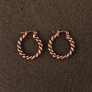 May include: A pair of rose gold hoop earrings featuring a twisted rope texture. Each circular earring is constructed with a hinged post mechanism at the top, showcasing a metallic finish that reflects light across the spiraled surface design.