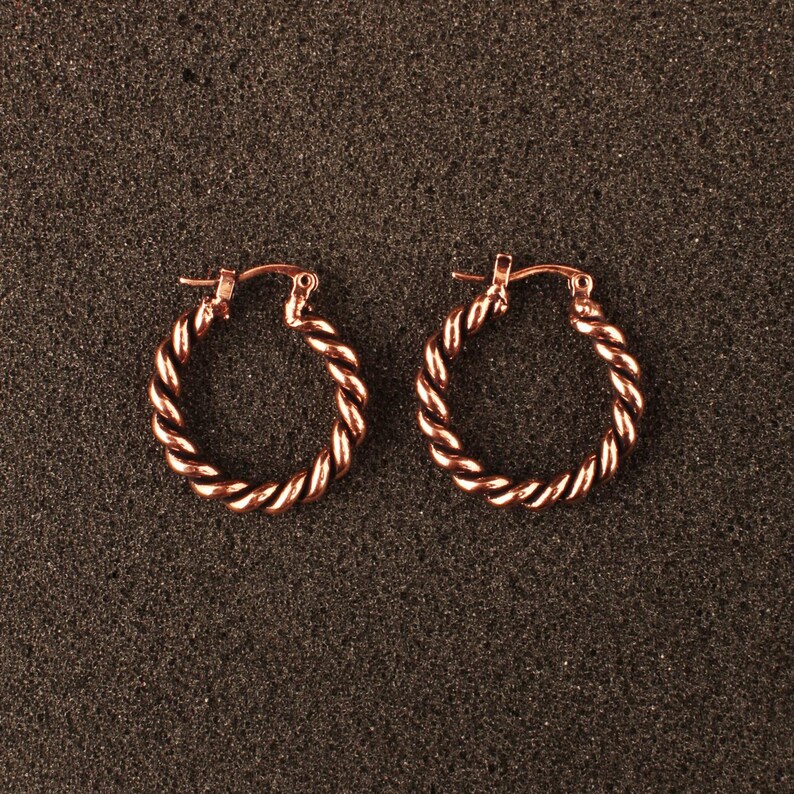 May include: A pair of rose gold hoop earrings featuring a twisted rope texture. Each circular earring is constructed with a hinged post mechanism at the top, showcasing a metallic finish that reflects light across the spiraled surface design.