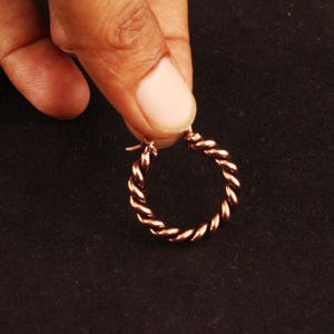 May include: A small, circular hoop earring crafted from polished copper-toned metal. The earring features a distinct twisted rope texture throughout its entire circumference, with a simple hinged closure mechanism visible at the top of the hoop.