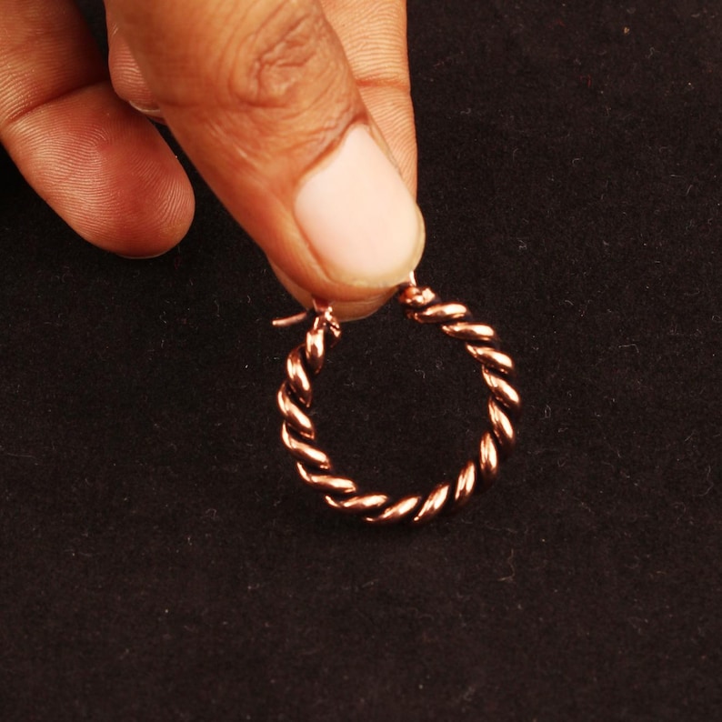 May include: A small, circular hoop earring crafted from polished copper-toned metal. The earring features a distinct twisted rope texture throughout its entire circumference, with a simple hinged closure mechanism visible at the top of the hoop.