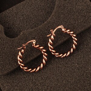 May include: A pair of rose gold hoop earrings featuring a twisted rope texture. Each circular earring is constructed with a hinged post mechanism for secure closure, showcasing a polished metallic finish that reflects light across the spiral design.