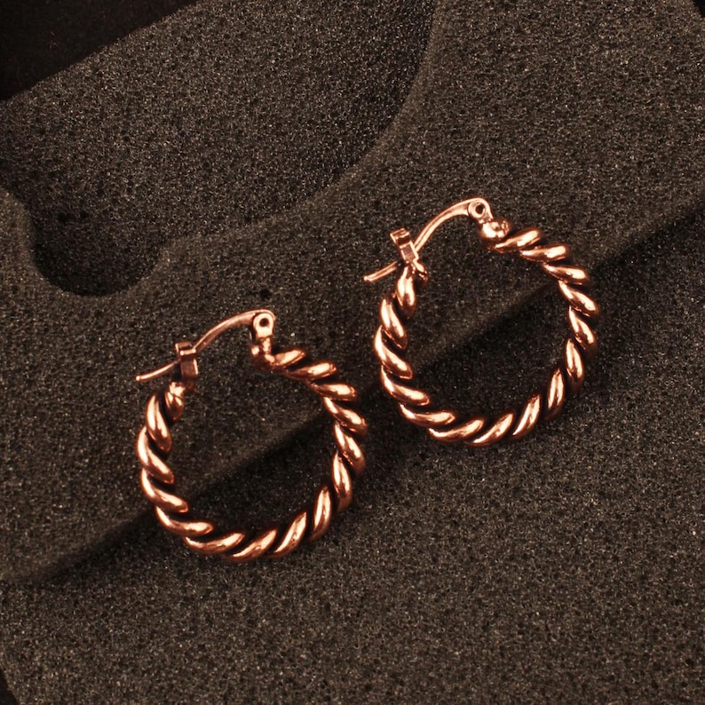 May include: A pair of rose gold hoop earrings featuring a twisted rope texture. Each circular earring is constructed with a hinged post mechanism for secure closure, showcasing a polished metallic finish that reflects light across the spiral design.