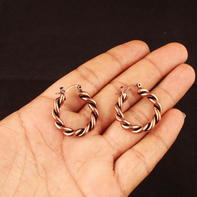 May include: A pair of small hoop earrings crafted from copper-toned metal. Each earring features a continuous twisted rope design with a smooth, polished finish and a hinged post closure mechanism that secures the circular hoop shape.