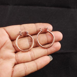 May include: A pair of small, circular hoop earrings with a polished copper finish. Each earring features a simple, continuous round band design with a hinged post closure mechanism at the top, showcasing a warm, metallic reddish-brown tone.
