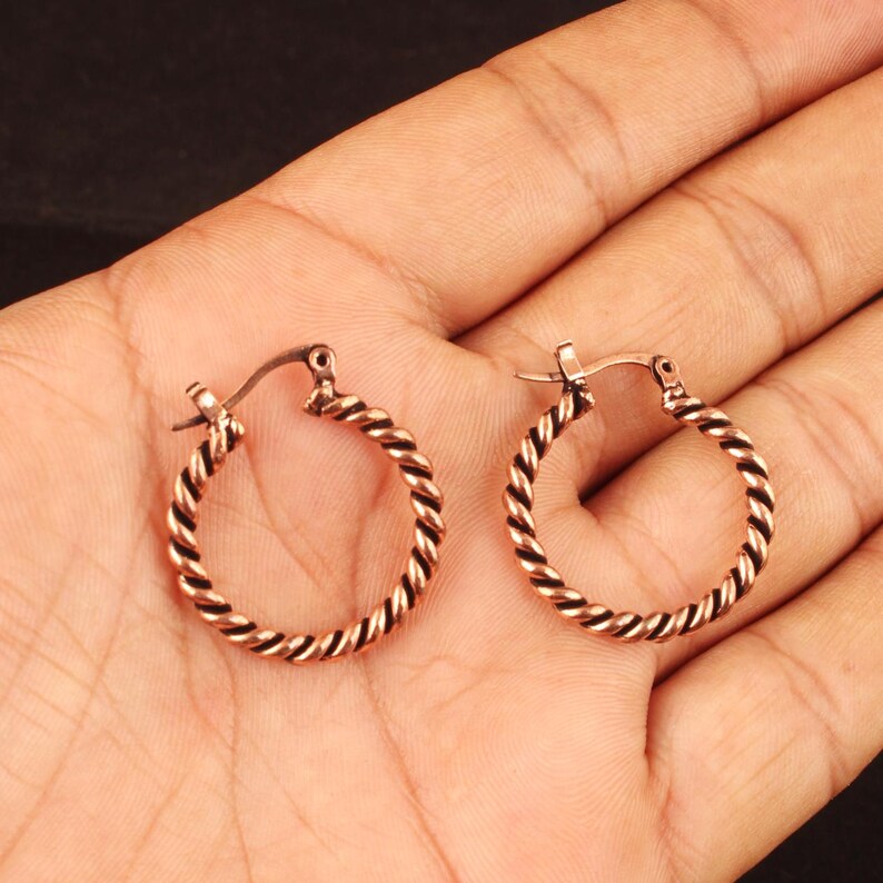 May include: A pair of small hoop earrings featuring a twisted rope texture. The earrings are crafted from a metallic material with a warm, copper-toned finish and include a hinged post closure mechanism for securing the hoops in place.