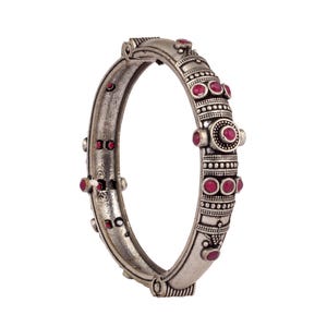 Puede incluir: Un brazalete rígido articulado de metal plateado oxidado, con una superficie texturizada con abalorios intrincados y pequeñas piedras cabujón redondas de color rosa intenso engastadas a intervalos regulares a lo largo de los bordes.