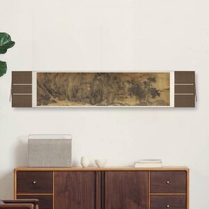 May include: A black and white ink painting of a landscape scene with mountains, trees, and figures. The painting is mounted on a scroll and displayed on a wall in a modern living room setting.