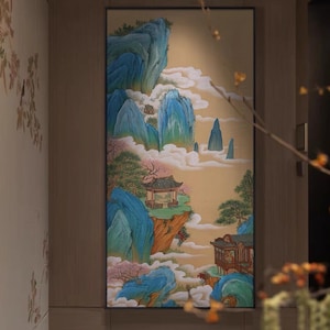 Könnte beinhalten: Gerahmtes Landschaftsgemälde mit einer traditionellen asiatischen Berglandschaft. Das Kunstwerk zeigt blaue Berge, weiße Wolken und kleine Gebäude mit roten Dächern. Die Farbpalette umfasst Blau-, Grün- und Goldtöne.