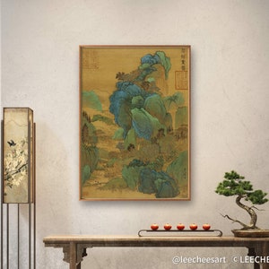 May include: A framed painting of a landscape with green and blue trees and mountains. The painting is on a gold background and has Chinese characters in the upper right corner. The painting is hanging on a white wall above a wooden table with a bonsai tree on the right side.