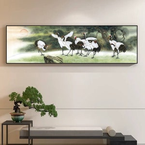 May include: A black framed painting depicting a group of white cranes with black wing tips and red accents on their heads. The cranes are standing in a green grassy field with a background of trees and a hazy sky.