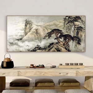 May include: A framed painting of a traditional Chinese landscape with mountains, trees, and clouds. The painting is in shades of gray, brown, and white, and is set against a white wall. The painting is hanging above a wooden table with a teapot and cups.