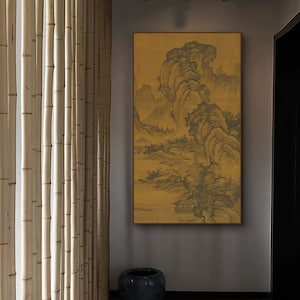 May include: A framed landscape painting with a gold background and a mountain scene. The painting is mounted on a gray wall, with a dark blue ceramic pot below. Bamboo poles are visible to the left.