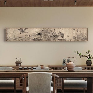 May include: A long, horizontal black and white ink painting of birds and reeds on a beige background. The painting is framed in a thin, brown frame and hangs on a beige wall above a wooden table with chairs.