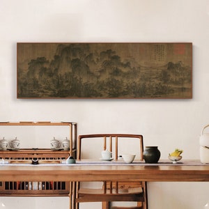 Pode incluir: Uma pintura de paisagem em preto e branco emoldurada que representa uma cordilheira com árvores e um rio. A pintura está pendurada em uma parede branca acima de uma mesa de madeira com xícaras de chá e um vaso.