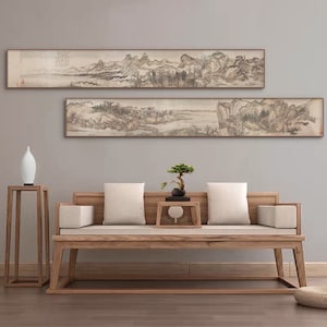 以下が含まれることがあります： 山、木、水のある風景を描いた、額縁に入った白黒の墨絵が2枚。絵は薄い灰色の壁に、木製のソファと白いクッションの上に飾られています。