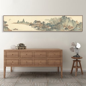 May include: A long, horizontal painting depicting a traditional Chinese landscape scene with mountains, trees, and a river. The painting is framed in a brown wood frame and hangs on a white wall above a wooden dresser.