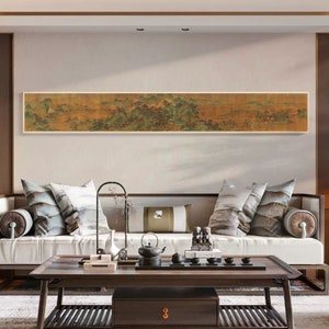 May include: A long, horizontal painting depicting a landscape scene with mountains, trees, and a river. The painting is framed in a simple, wooden frame and is hanging on a wall in a modern living room. The painting is a traditional Chinese landscape painting, with a focus on the natural world and the beauty of the scenery.