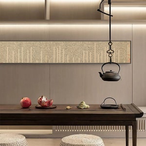 May include: A dark wood table with two woven stools, a black teapot hanging from the ceiling, and two pomegranates on a plate. The table is set with a black tray and a woven mat. The background is a light beige wall with a long scroll hanging on the wall.