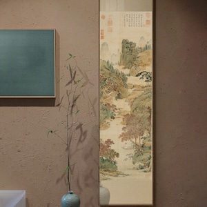 May include: A framed vertical painting of a traditional Chinese landscape scene with mountains, trees, and a river. The painting is in shades of brown, green, and white, with a few red accents. The painting is framed in a thin, gold-colored frame.