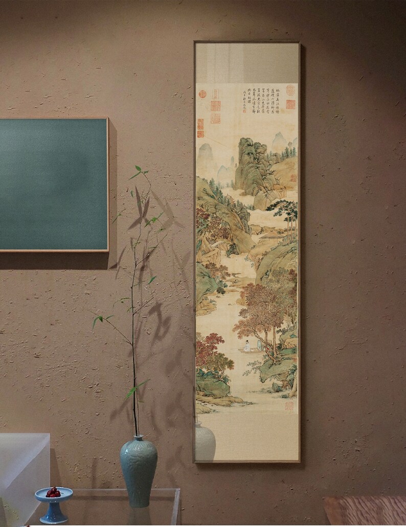 May include: A framed vertical painting of a traditional Chinese landscape scene with mountains, trees, and a river. The painting is in shades of brown, green, and white, with a few red accents. The painting is framed in a thin, gold-colored frame.