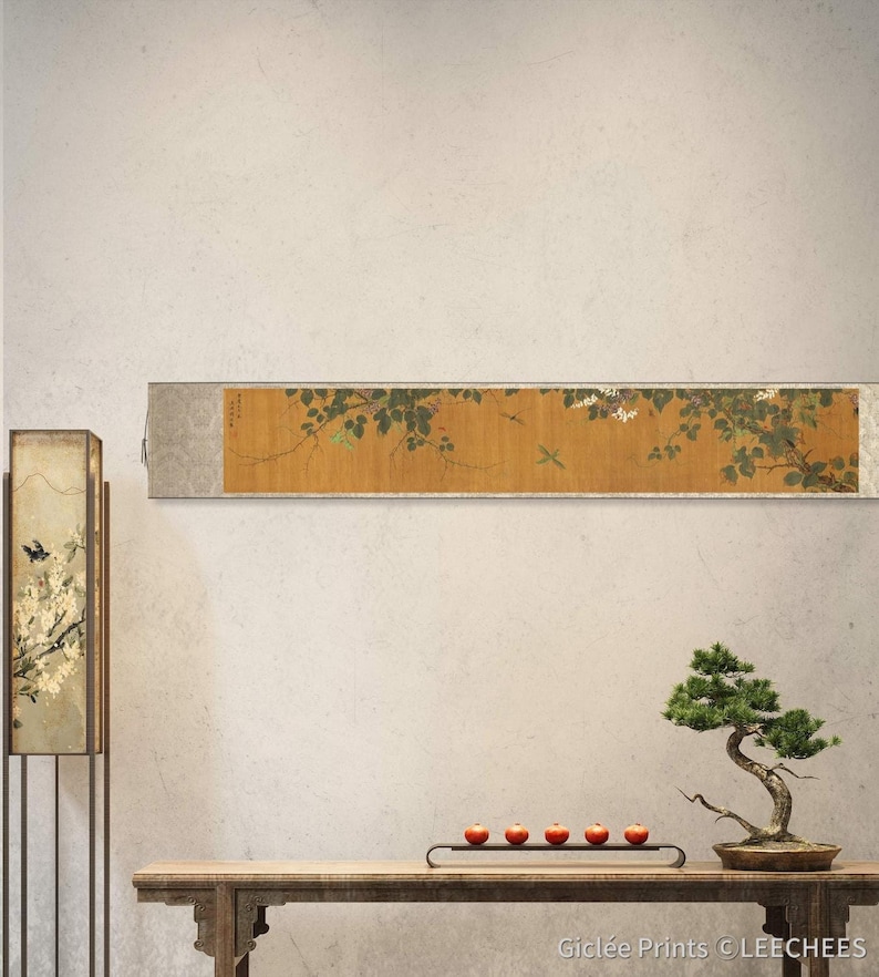 May include: A traditional Japanese scroll painting depicting a branch of a flowering tree with delicate white blossoms and green leaves. The scroll is mounted on a wall in a modern interior with a wooden table, a bonsai tree, and a lamp.