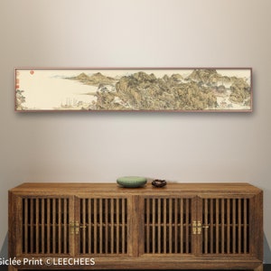 May include: A long, horizontal painting of a landscape with mountains, trees, and water. The painting is framed in a brown wood frame and is hanging on a white wall above a wooden cabinet with two doors.