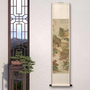 May include: A traditional Chinese scroll painting depicting a landscape scene with mountains, trees, and a river. The painting is done in shades of brown, green, and white, and features a small figure in a boat on the river. The scroll is mounted on a wooden dowel and has a decorative tassel.