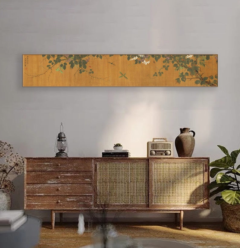 May include: A long, horizontal painting depicting a branch of a tree with green leaves and white flowers. The painting is mounted on a wall above a wooden cabinet with a woven rattan door.