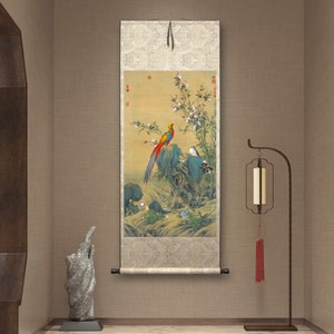 Op de afbeelding: Een traditioneel Chinees schilderij op een rol, waarop een kleurrijke fazant te zien is die op een rots zit met bloeiende bloemen en een kleine vogel. De rol is gemonteerd op een houten stok en hangt aan een beige muur.