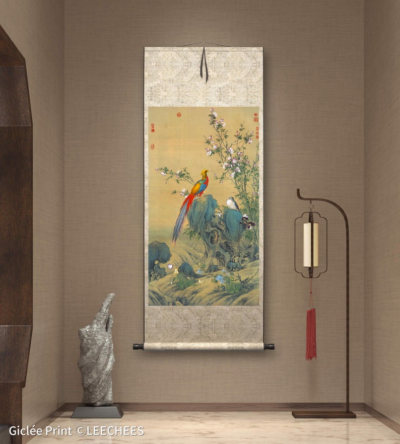 Op de afbeelding: Een traditioneel Chinees schilderij op een rol, waarop een kleurrijke fazant te zien is die op een rots zit met bloeiende bloemen en een kleine vogel. De rol is gemonteerd op een houten stok en hangt aan een beige muur.