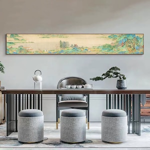 May include: A long, horizontal painting depicting a traditional Chinese landscape scene with mountains, trees, water, and a small village. The painting is framed and hanging on a wall above a wooden table with two grey fabric ottomans and a black chair.