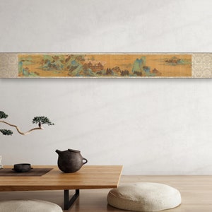 May include: A long, horizontal scroll painting depicting a landscape scene with mountains, trees, and a building. The painting is on a light yellow background and is framed in a dark wood frame.