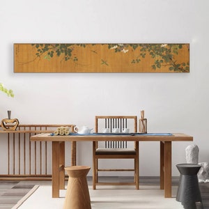 May include: A long, horizontal painting of a vine with white flowers and green leaves. The painting is framed in a thin, wooden frame and is hanging on a white wall above a wooden table and chairs.
