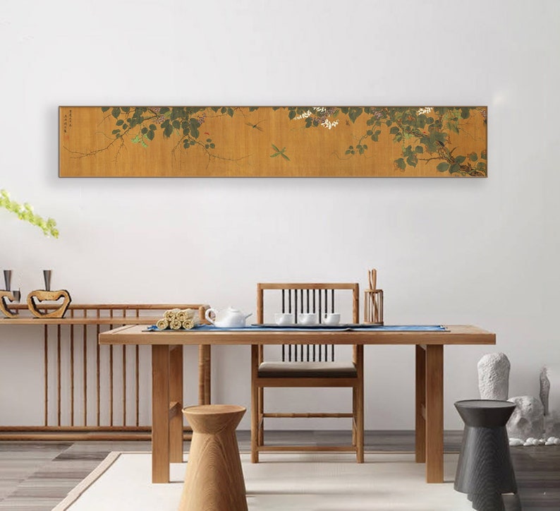 May include: A long, horizontal painting of a vine with white flowers and green leaves. The painting is framed in a thin, wooden frame and is hanging on a white wall above a wooden table and chairs.