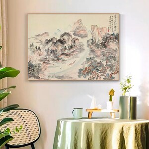 May include: A traditional Chinese ink and wash painting depicting a landscape scene with mountains, trees, and a river. The painting is done in shades of grey, black, and white, with hints of pink and blue. The painting is framed in a light wood frame.