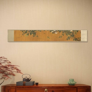 May include: A framed scroll painting depicting a branch of a flowering tree with delicate white blossoms and green leaves. Two small birds are perched on the branches. The scroll is mounted on a beige wall above a wooden table with a teapot, a small box, and a vase.