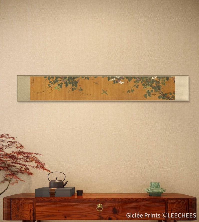 May include: A framed scroll painting depicting a branch of a flowering tree with delicate white blossoms and green leaves. Two small birds are perched on the branches. The scroll is mounted on a beige wall above a wooden table with a teapot, a small box, and a vase.
