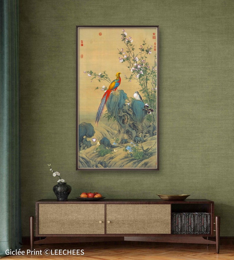 Op de afbeelding: Een ingelijst schilderij van een kleurrijke fazant die op een rots zit, omringd door witte bloemen en andere vogels. Het schilderij hangt aan een groene muur, met daaronder een houten kast en een vaas met bloemen.