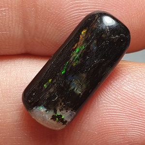 May include: A polished, rectangular black opal gemstone. The opal displays iridescent flashes of green, orange, and blue within its dark surface. The stone has a rounded top and a lighter, textured base. The opal is held in a person's hand.
