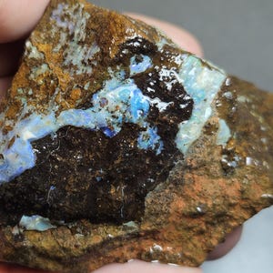 Könnte beinhalten: Ein rohes Opal-Exemplar mit einer Mischung aus Farben. Der Stein weist Bereiche in leuchtendem Blau, Schwarz und Braun auf, mit einer strukturierten Oberfläche. Der Opal wird in einer Hand gehalten, was seine natürliche, rohe Form zeigt.