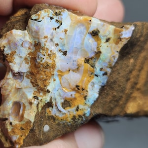 Könnte beinhalten: Ein rohes Opal-Exemplar mit einer Mischung aus Farben. Der Opal weist Bereiche in Weiß, Blau und Gelb mit brauner Matrix auf. Der Stein hat eine strukturierte Oberfläche und eine unregelmäßige Form, die natürliche Muster und Variationen zeigt.