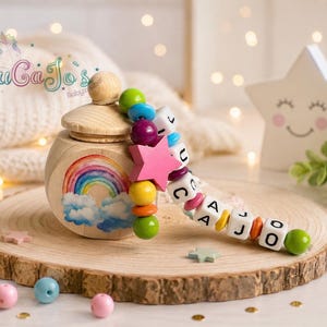 Könnte beinhalten: Eine Holzkiste mit Regenbogen- und Wolkenmuster, gekrönt von einem Holzdeckel und einem Perlenzubehör. Das Zubehör verfügt über bunte Perlen, einen rosa Stern und Buchstabenwürfel, die einen Namen buchstabieren. Die Box steht auf einer Holzscheibe.