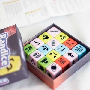 Op de afbeelding: Bordspel "Randice" met een doos en dobbelstenen. De dobbelstenen zijn vierkant en tonen symbolen en woorden zoals "HUMAN" en "SIM". De doos is donker met de naam van het spel en een geel dollarteken.