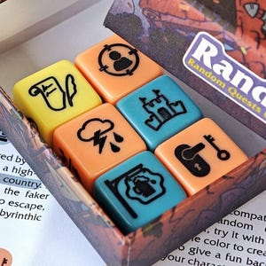 Puede incluir: Una caja de juego de mesa etiquetada como "Randice: Random Quests Generator" con dados coloridos. Los dados son amarillos, naranjas y azules, cada uno con un icono negro único. La caja tiene una ilustración detallada.