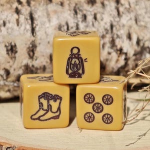 Puede incluir: Tres dados de seis caras de color amarillo claro con iconos grabados en marrón oscuro. Un dado muestra una linterna, otro un par de botas vaqueras y el tercer dado presenta un patrón de cinco pequeños símbolos circulares tipo rueda.