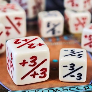 Pode incluir: Um conjunto de dados brancos de seis lados com marcações de frações matemáticas em vermelho e azul. O dado maior em primeiro plano exibe +3/3 e +2/2 em vermelho, enquanto o menor mostra -5/5 e -3/3 em azul, sobre uma superfície.