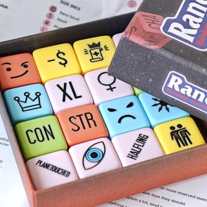 Puede incluir: Un juego de mesa con fichas cuadradas de colores en una caja. Las fichas presentan símbolos y palabras como "XL", "STR" y "PLANETOUCHED". La caja está etiquetada como "Randice" y "Random Character Generator".