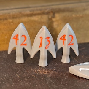 Puede incluir: Tres piezas de juego grises en forma de flecha con números naranjas: 42, 13 y 42. Las piezas están sobre una superficie marrón. Hay piezas adicionales visibles en el fondo. Las puntas de flecha son blancas.