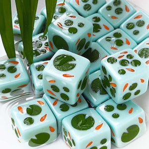 May include: A collection of light blue dice featuring a pond-themed design with dark green lily pads and small orange koi fish patterns on each face. The dice have a smooth, glossy finish and are arranged in a cluster with green plant leaves.